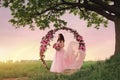 Beautiful bride in pink dress standing with her bouquet at a round flower arch under a large single tree in a field at sunset.