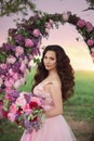 Beautiful bride in pink dress standing with her bouquet at a round flower arch under a large single tree in a field at sunset. Royalty Free Stock Photo