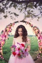 Beautiful bride in pink dress standing with her bouquet at a round flower arch under a large single tree in a field at sunset. Royalty Free Stock Photo