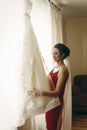 Beautiful bride in red robe with veil looking at elegant white lace wedding dress on hanger near a window, morning wedding Royalty Free Stock Photo