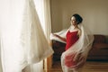 Beautiful bride in red robe with veil looking at elegant white lace wedding dress on hanger near a window, morning wedding Royalty Free Stock Photo