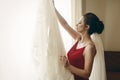 Beautiful bride in red robe with veil looking at elegant white lace wedding dress on hanger near a window, morning wedding Royalty Free Stock Photo