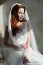 Beautiful redhair lady in elegant white wedding dress. Fashion portrait of model indoors. Beauty woman sitting near her bed Royalty Free Stock Photo