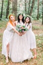 Beautiful bride in stylish rustic dress and wreath is holding flower bouquet in her hands. Two pretty bridesmaids in