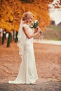 Beautiful bride in white long dress is smelling wedding bouquet in autumn park on wedding day