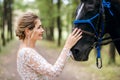 Beautiful bride in a white wedding dress stroking a horse, portrait of a blonde woman with a black stallion, wedding day Royalty Free Stock Photo