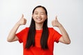 Beautiful brunette asian girl shows her suppport, winks and shows thumbs up in approval, like something, agree and Royalty Free Stock Photo