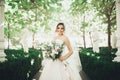 Beautiful brunette bride in elegant white dress holding bouquet posing neat trees