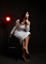 Beautiful brunette business woman in stylish white elegant dress barefoot sitting on stool looking away over dark