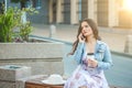 Beautiful brunette girl in a jeans jacket talking on the phone sitting on a bench in the city and holding a coffee Royalty Free Stock Photo