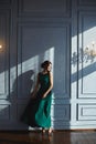 A beautiful brunette girl with modish hairstyle, in green evening dress, dancing near the blue wall with sunlight and