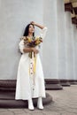 A beautiful brunette girl in a white fashionable dress with a bouquet of wild wildflowers posing near the stone columns Royalty Free Stock Photo