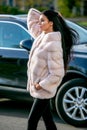 Beautiful brunette in a light-colored fur coat and black pants walks down the street in front of the car on an autumn sunny day Royalty Free Stock Photo