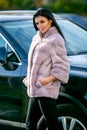 A beautiful brunette in a light-colored fur coat and black trousers is standing near a car on an autumn sunny day and is smiling s Royalty Free Stock Photo