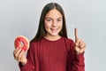 Beautiful brunette little girl holding tasty colorful doughnut smiling with an idea or question pointing finger with happy face, Royalty Free Stock Photo