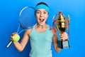 Beautiful brunette little girl playing tennis holding trophy celebrating crazy and amazed for success with open eyes screaming Royalty Free Stock Photo