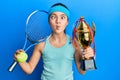 Beautiful brunette little girl playing tennis holding trophy making fish face with mouth and squinting eyes, crazy and comical Royalty Free Stock Photo