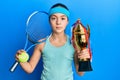 Beautiful brunette little girl playing tennis holding trophy relaxed with serious expression on face Royalty Free Stock Photo