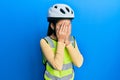 Beautiful brunette little girl wearing bike helmet and reflective vest with sad expression covering face with hands while crying