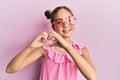 Beautiful brunette little girl wearing heart shaped sunglasses smiling in love doing heart symbol shape with hands Royalty Free Stock Photo