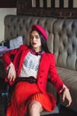 Beautiful brunette model with blue eyes in retro clothes: pink takes, white blouse, red jacket and skirt indoors