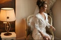 Beautiful brunette model girl with bright makeup in white dressing gown sits on the chair and posing at the interior Royalty Free Stock Photo