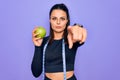 Beautiful brunette sporty woman using tape measure eating apple fruit over purple background pointing with finger to the camera Royalty Free Stock Photo