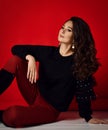 Beautiful brunette woman with curly hair in stylish clothing and accessories sitting on floor looking away