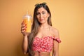 Beautiful brunette woman drinking glass with healthy orange juice over yellow background with a confident expression on smart face Royalty Free Stock Photo