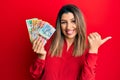 Beautiful brunette woman holding australian dollars pointing to the back behind with hand and thumbs up, smiling confident Royalty Free Stock Photo