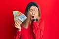 Beautiful brunette woman holding australian dollars yawning tired covering half face, eye and mouth with hand