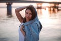 a beautiful brunette woman poses with a denim jacket in her hands over her head at sunset and a bridge over the river Royalty Free Stock Photo