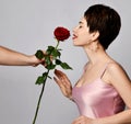 Beautiful brunette woman with short hair in pink silk dress and earrings standing and sniffing red rose flower in mans hand