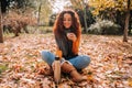 A beautiful brunette woman sitting on the ground surrounded by leaves while enjoying a coffee in the park. She is putting the