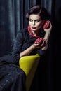 Beautiful brunette woman wearing black evening dress posing with two pomegranates in hands while sitting on yellow chair Royalty Free Stock Photo