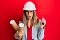 Beautiful brunette woman wearing safety helmet holding blueprints in shock face, looking skeptical and sarcastic, surprised with Royalty Free Stock Photo