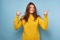 A beautiful brunette in a yellow sweater stands on a blue background, smiling embarrassed, raising her thumbs to the top