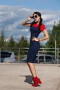 Beautiful brunette young woman in blue and red dress, wearing red high-heeled shoes, sunglasses, walking along the street. Style o Royalty Free Stock Photo