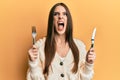 Beautiful brunette young woman holding fork and knife ready to eat angry and mad screaming frustrated and furious, shouting with