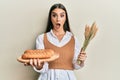 Beautiful brunette young woman holding homemade bread and spike wheat afraid and shocked with surprise and amazed expression, fear