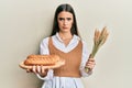 Beautiful brunette young woman holding homemade bread and spike wheat skeptic and nervous, frowning upset because of problem