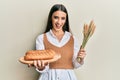 Beautiful brunette young woman holding homemade bread and spike wheat smiling and laughing hard out loud because funny crazy joke