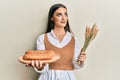 Beautiful brunette young woman holding homemade bread and spike wheat smiling looking to the side and staring away thinking