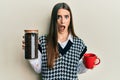 Beautiful brunette young woman holding jar with coffee beans and drinking coffee cup in shock face, looking skeptical and Royalty Free Stock Photo