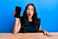 Beautiful brunette young woman holding smartphone showing blank screen scared and amazed with open mouth for surprise, disbelief Royalty Free Stock Photo