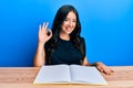 Beautiful brunette young woman sitting on desk with open blank book doing ok sign with fingers, smiling friendly gesturing Royalty Free Stock Photo
