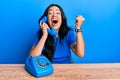 Beautiful brunette young woman speaking on vintage telephone screaming proud, celebrating victory and success very excited with Royalty Free Stock Photo