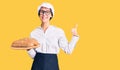 Beautiful brunette young woman wearing baker uniform holding homemade bread smiling happy and positive, thumb up doing excellent Royalty Free Stock Photo