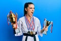 Beautiful brunette young woman wearing karate fighter uniform and medals holding trophy angry and mad screaming frustrated and