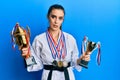 Beautiful brunette young woman wearing karate fighter uniform and medals holding trophy clueless and confused expression